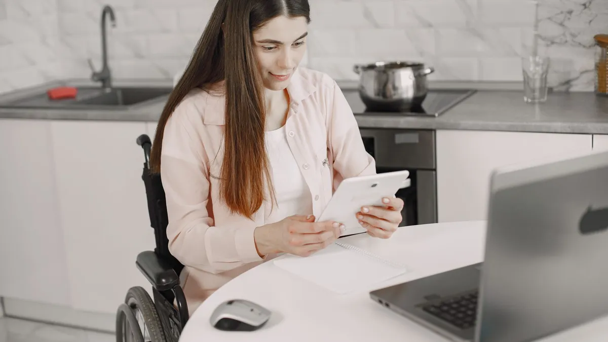 Woman in wheelchair engaged with tablet at kitchen table, creating an inclusive work environment.