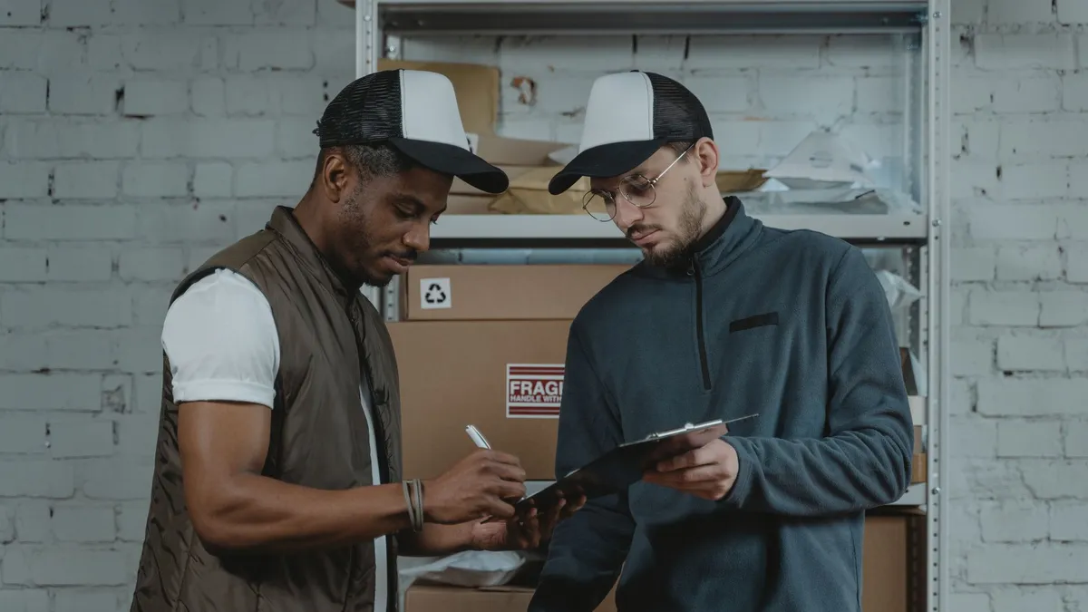 Two male couriers discussing and signing a clipboard in a warehouse setting, surrounded by packages.