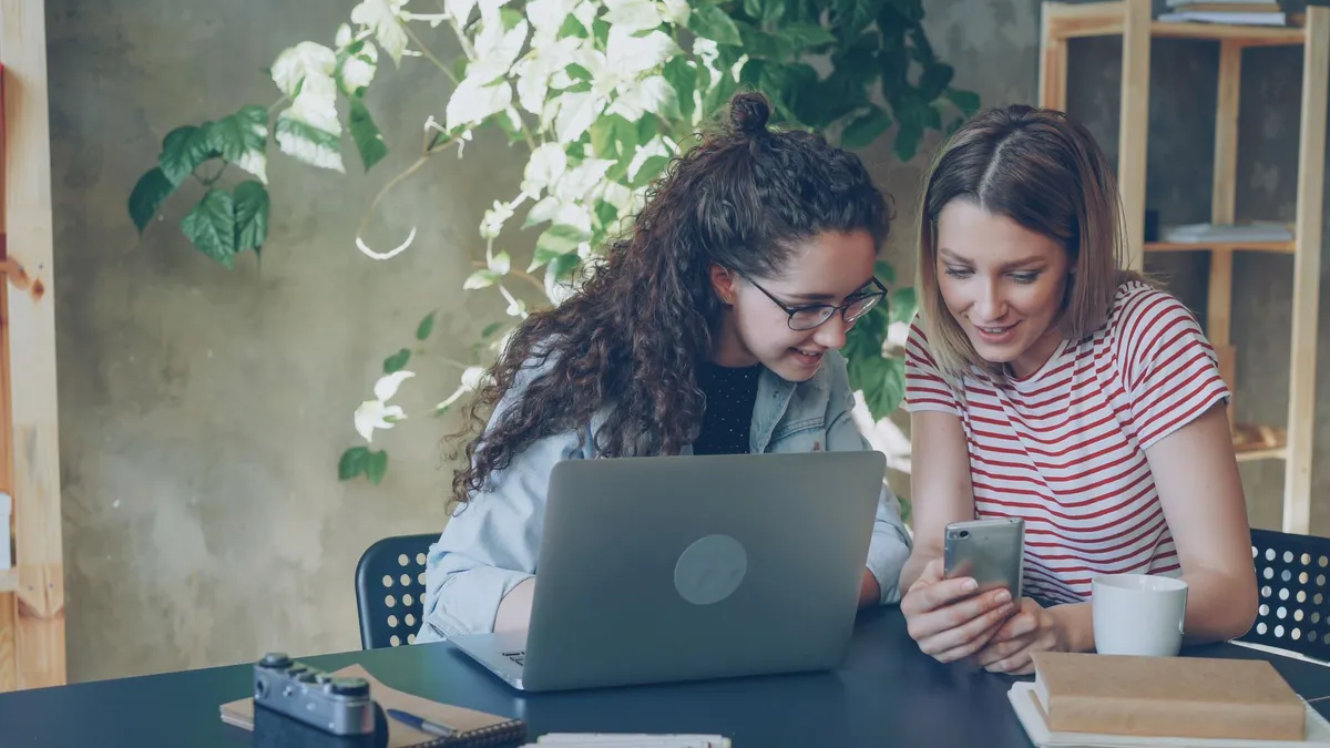 Two women browsing a smartphone while working on a project in a cozy indoor setting.