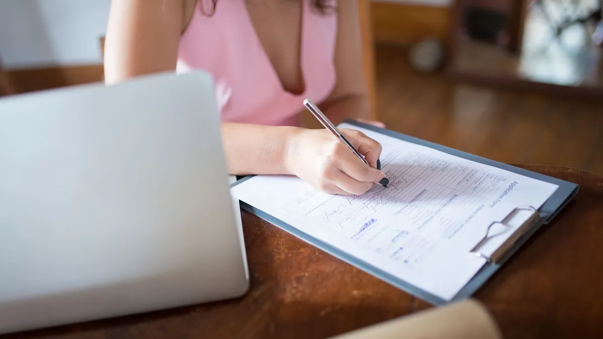 A woman writes on a document clipped to a clipboard, with a laptop nearby on a wooden desk.