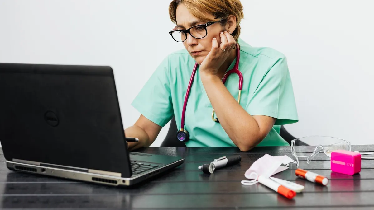 Focused nurse with eyeglasses working on a laptop surrounded by medical tools on a desk.