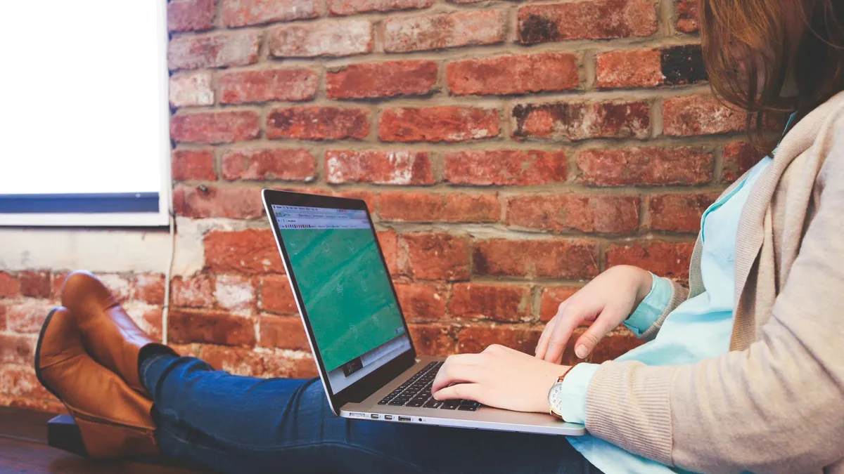 Woman working remotely on a laptop against a brick wall, representing modern flexible workspace.