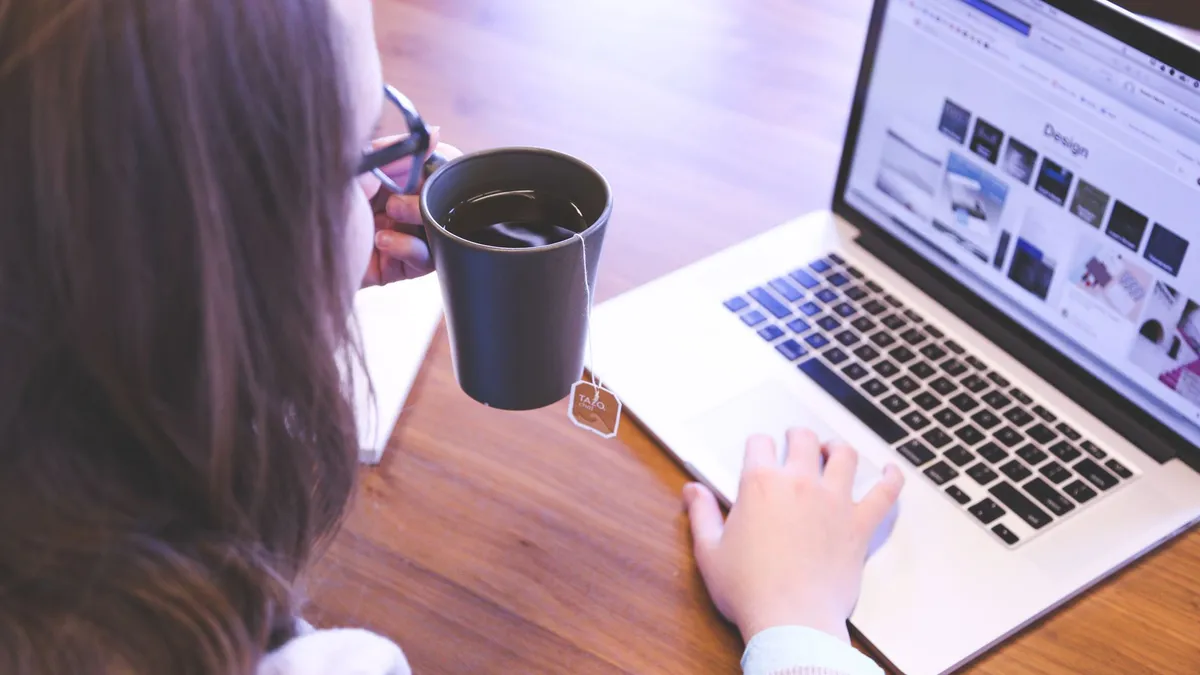 A woman enjoys coffee while browsing the web on a laptop at a home office desk.