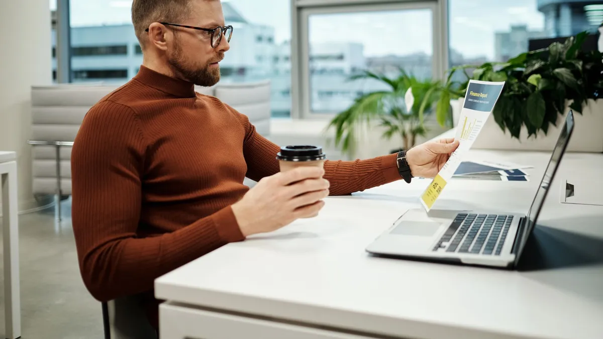 Professional businessman in brown turtleneck analyzing financial report while holding a takeaway coffee in modern office.