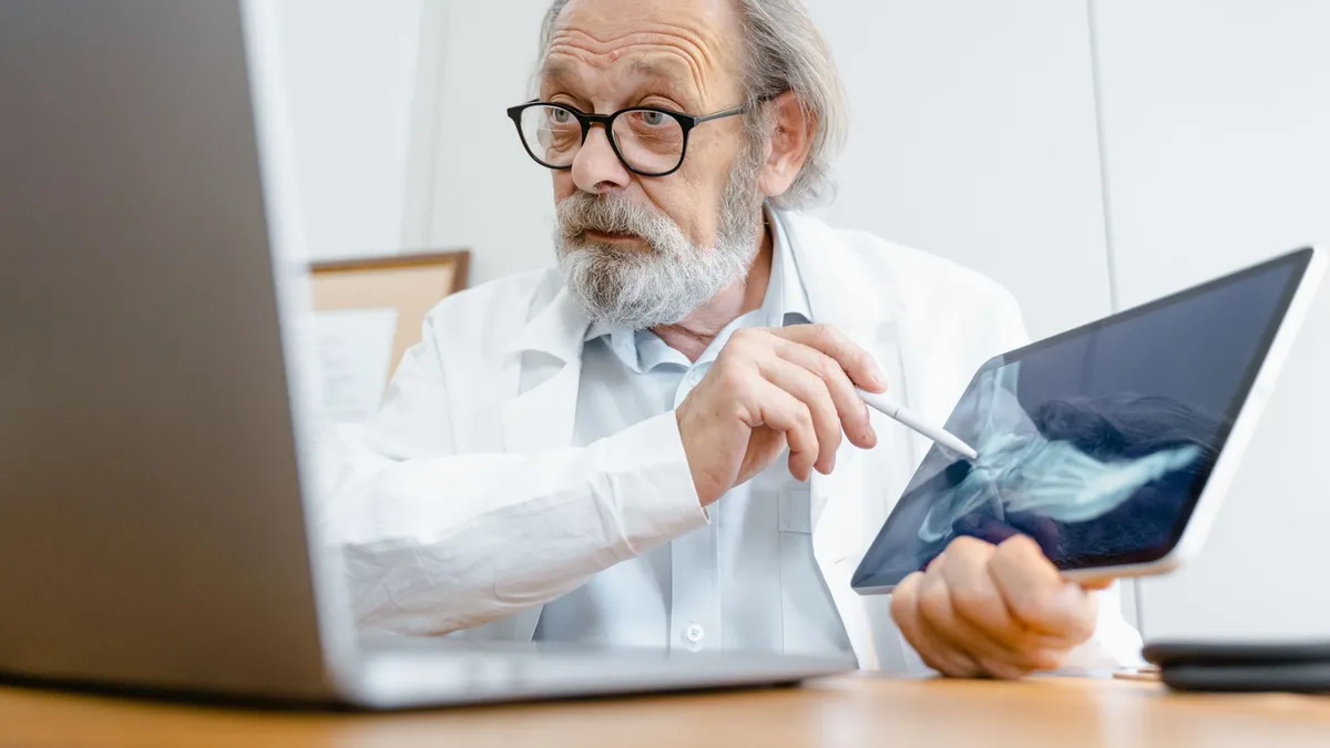 A senior doctor in a white coat explaining an X-ray result using a tablet in a medical office.