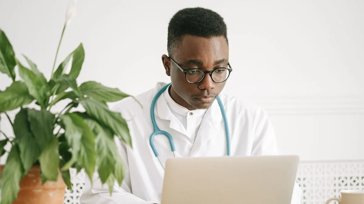 African American doctor working on a laptop in a bright office setting with potted plant.