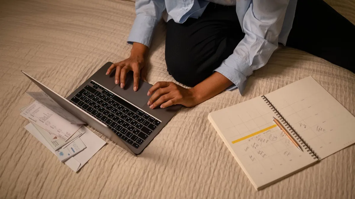 A woman working on a laptop while planning and organizing notes on a bed.