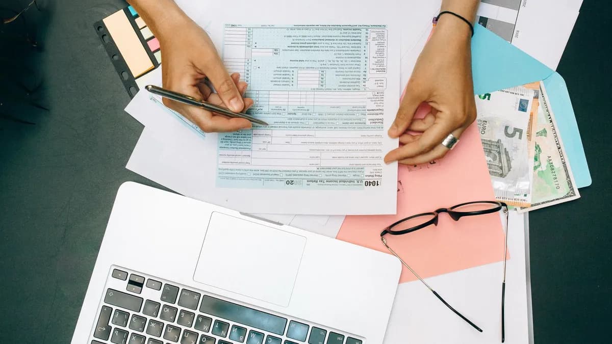 Hands writing on tax documents with laptop, glasses, and currency on desk.