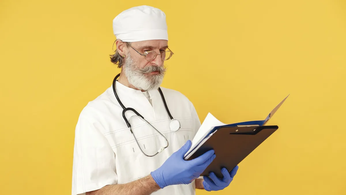 A bearded male doctor with stethoscope reviews medical documents on a yellow background.