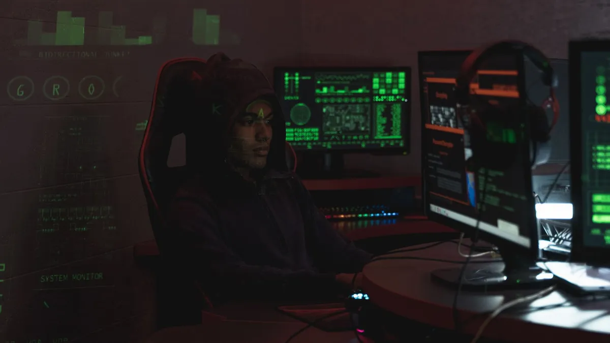 A man in a black hoodie engaged in cybersecurity work using multiple monitors indoors.