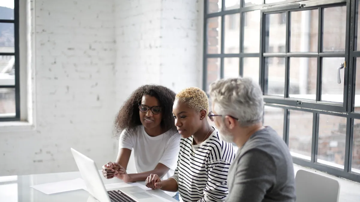 Side view of black women and old gray haired man sitting at table and surfing laptop while working on creative project