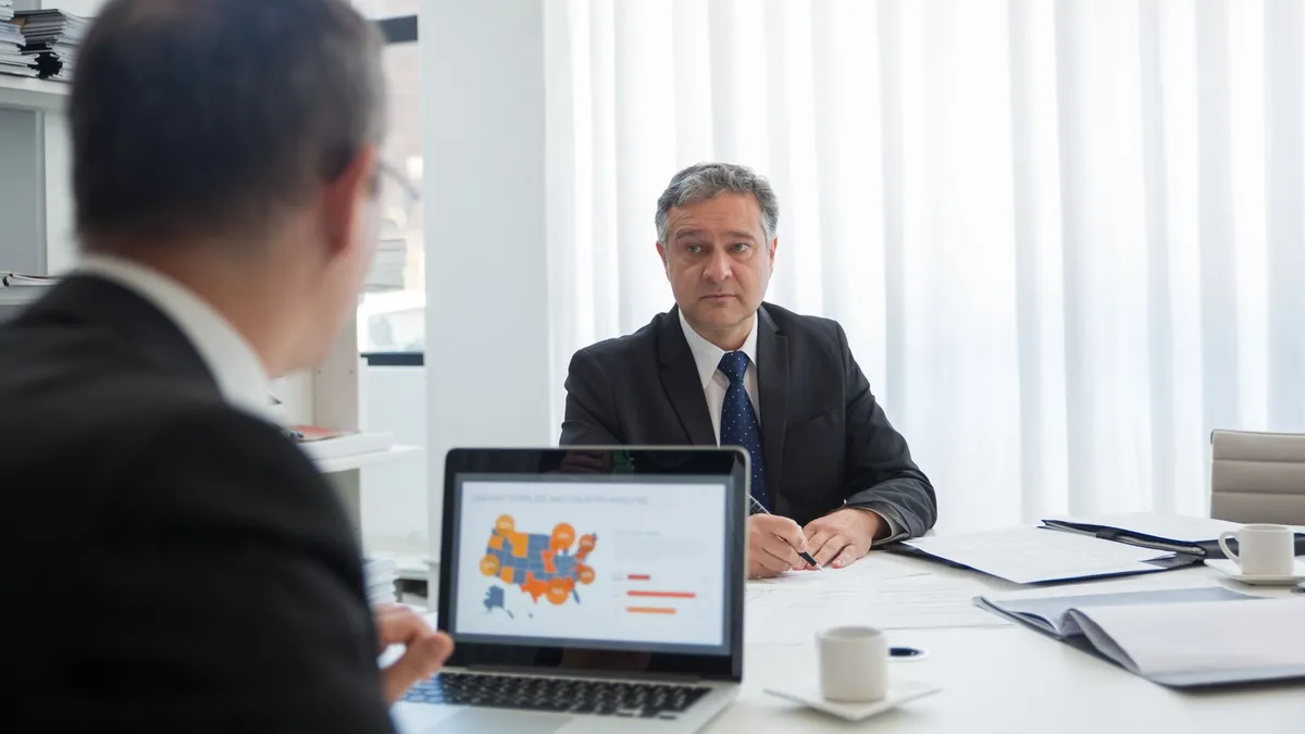 Two businessmen in discussion at an office desk with a laptop.