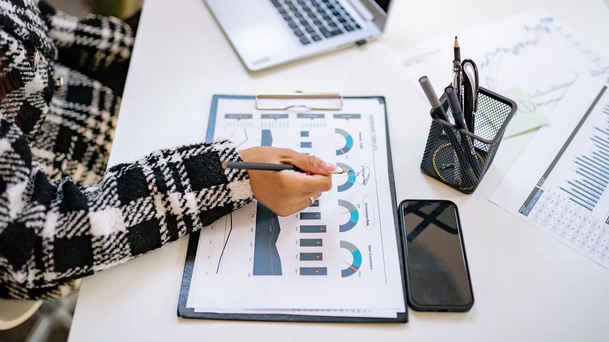 An office setting with a person analyzing charts and graphs at a desk with a laptop and smartphone.