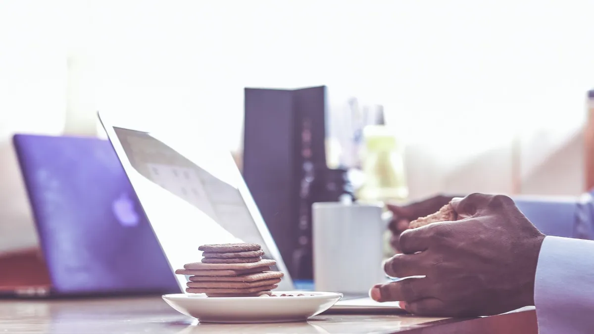 Hands holding coffee with biscuits on desk beside laptop during work break.