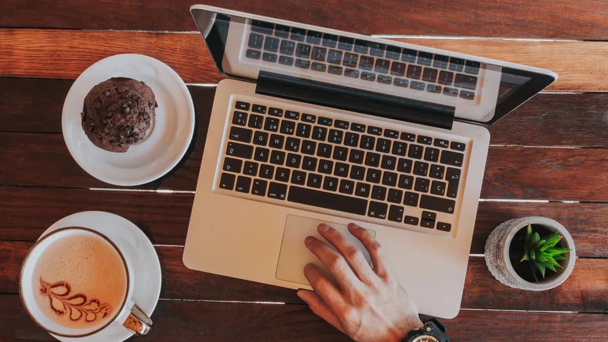 Overhead view of laptop, coffee cup with latte art, cookie, succulent, and hand on wooden table.