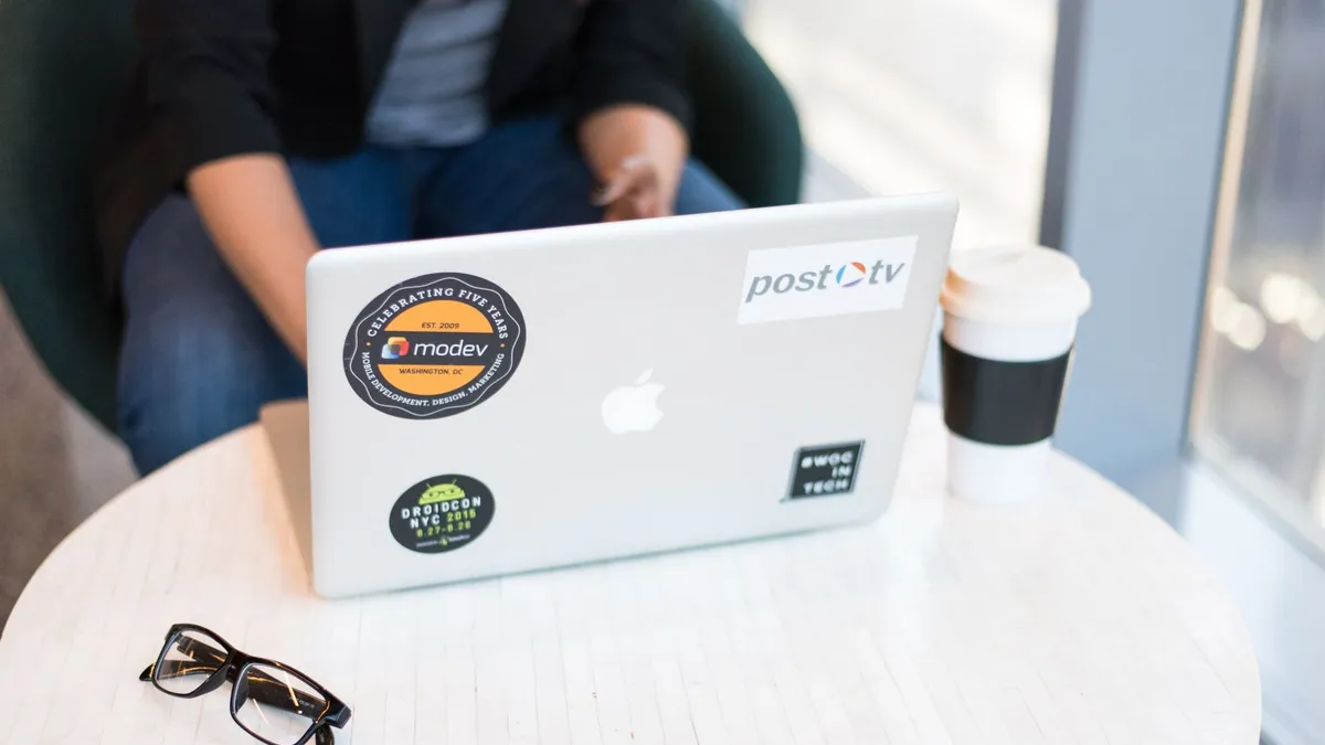 Person sitting indoors working on a laptop with stickers, coffee, and eyeglasses on a table.