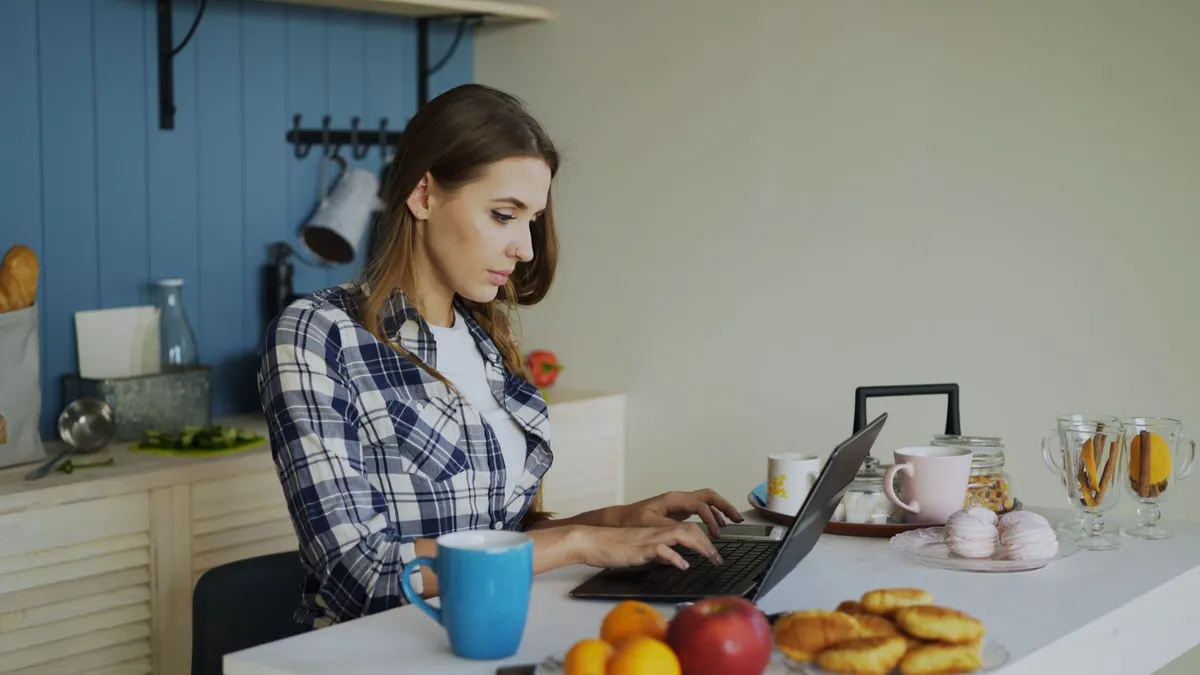 A woman using a laptop in a kitchen setting, surrounded by snacks and warm decor.