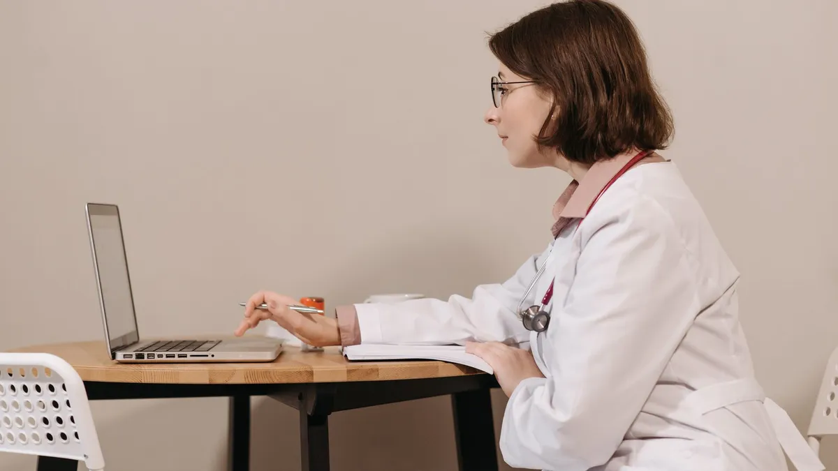 Side profile of a female doctor in a white coat working on a laptop at a desk.