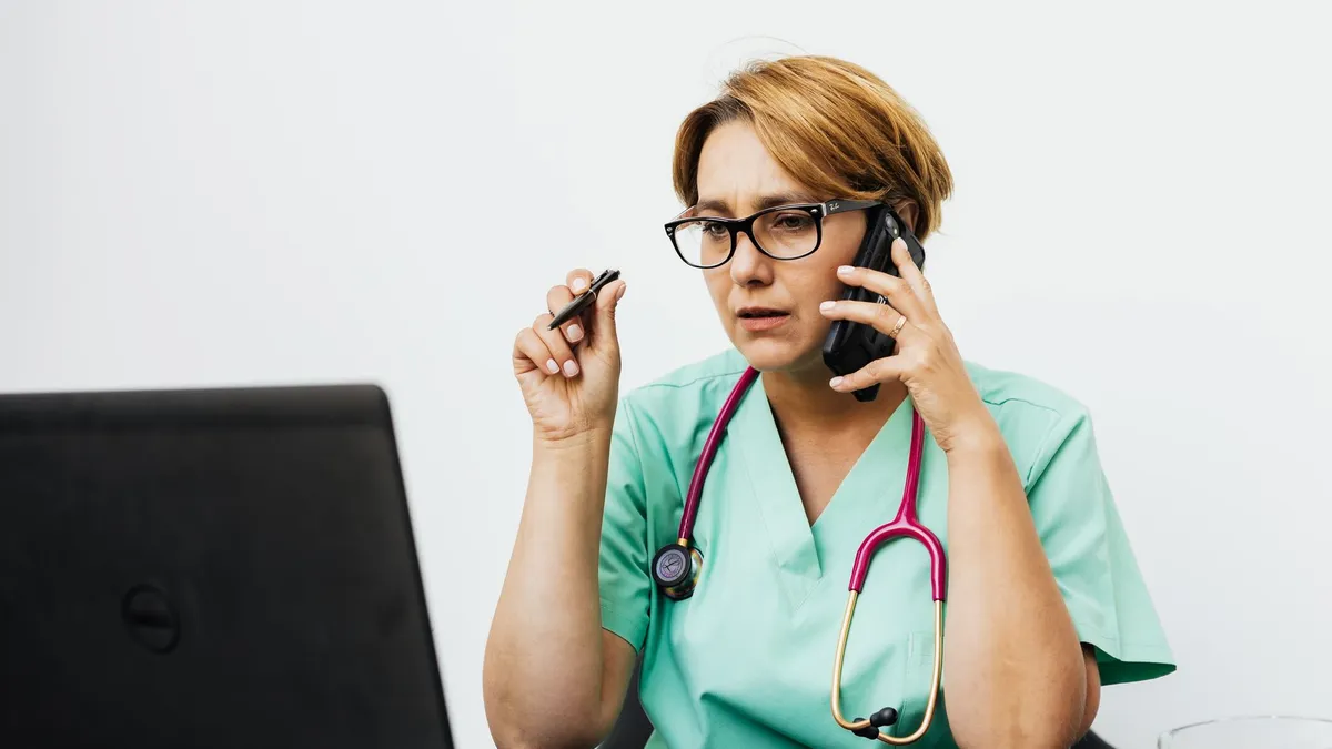 A woman in medical scrubs working with a laptop and phone, illustrating modern healthcare communication.