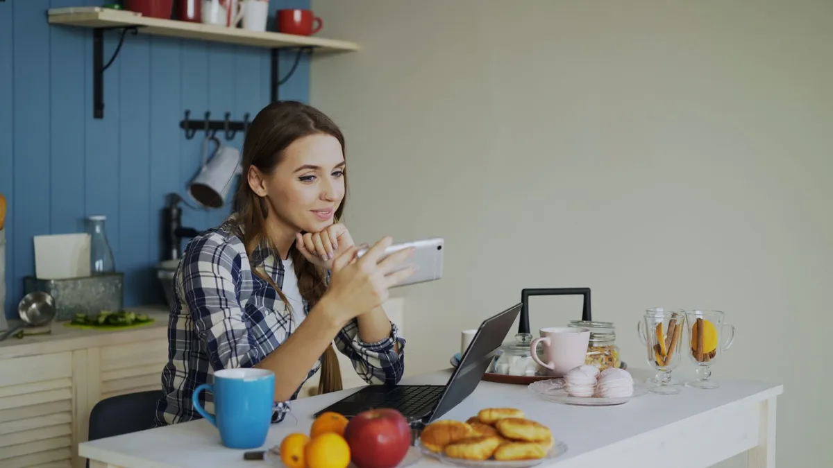 Young woman using smartphone and laptop while enjoying breakfast in cozy kitchen.