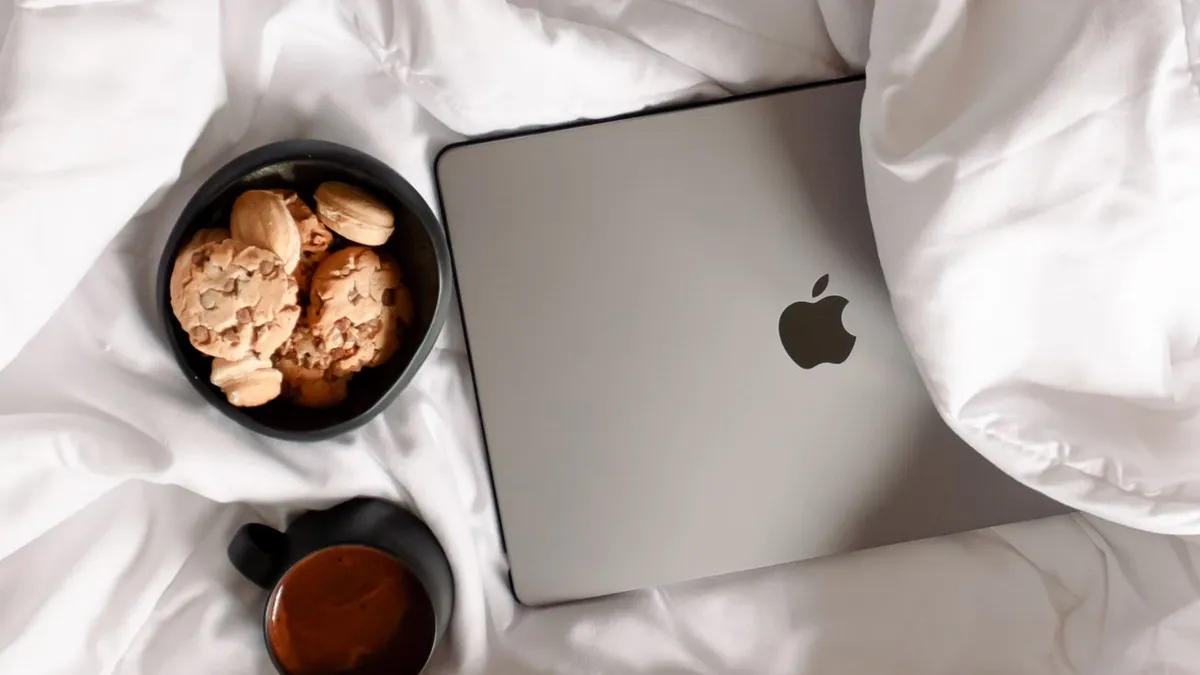 Flat lay of a laptop, coffee, and cookies on a comfortable bed setting.