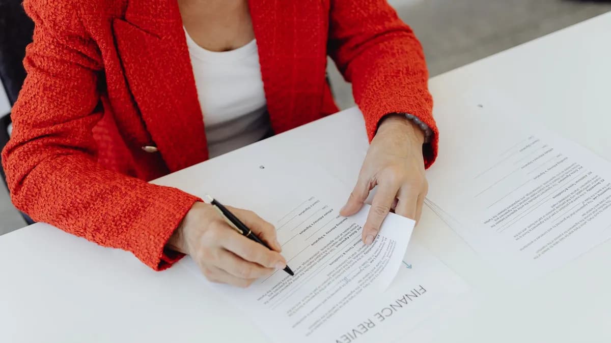 Close-up of a businesswoman in a red blazer signing finance documents at an office desk.