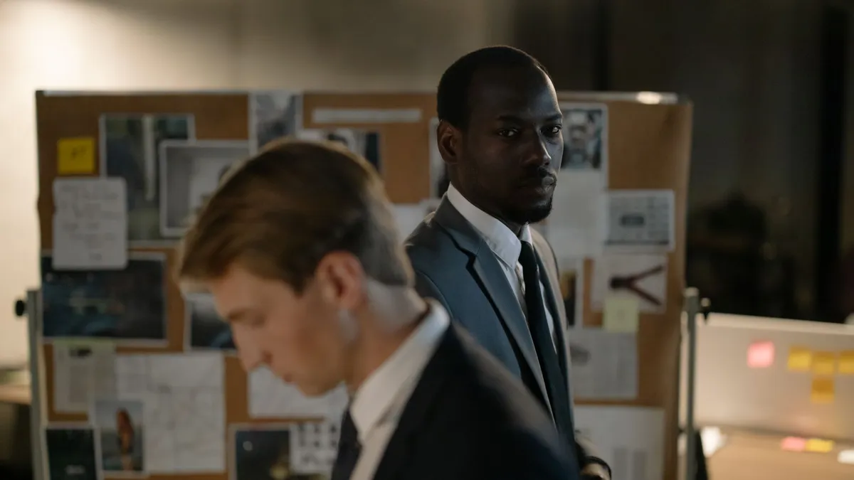 Two detectives examining a corkboard filled with evidence in a dimly lit office.