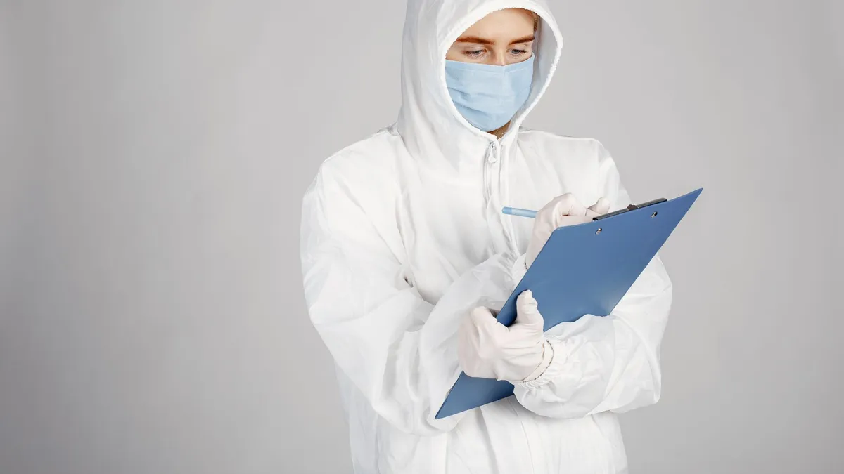 Healthcare professional in PPE suit writing on clipboard in a clinical setting.