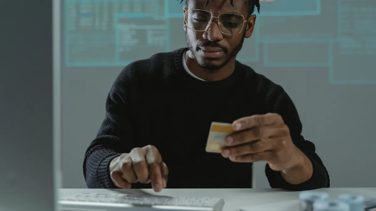 Man with glasses holding a bank card while typing on a keyboard, sitting in front of a computer.