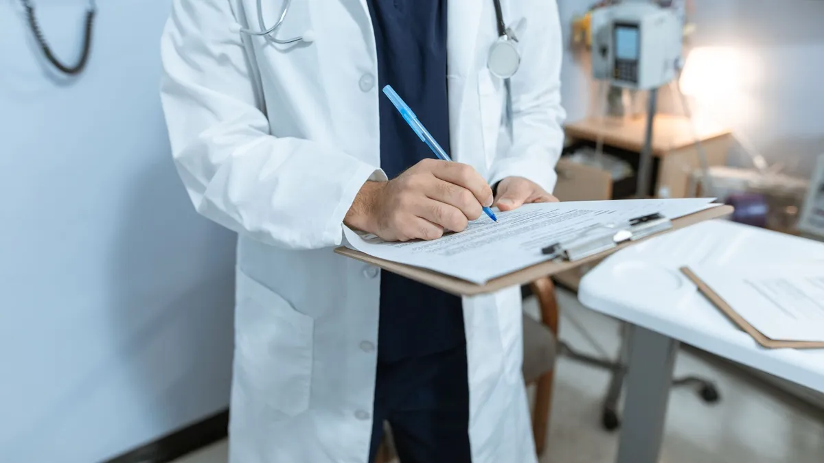 A doctor in a lab coat writing notes in a hospital setting, using a clipboard.