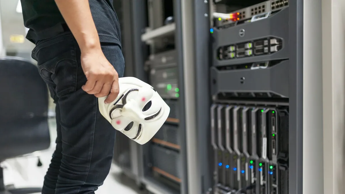 Person holding anonymous mask near servers, hinting at cybersecurity and hacking themes.
