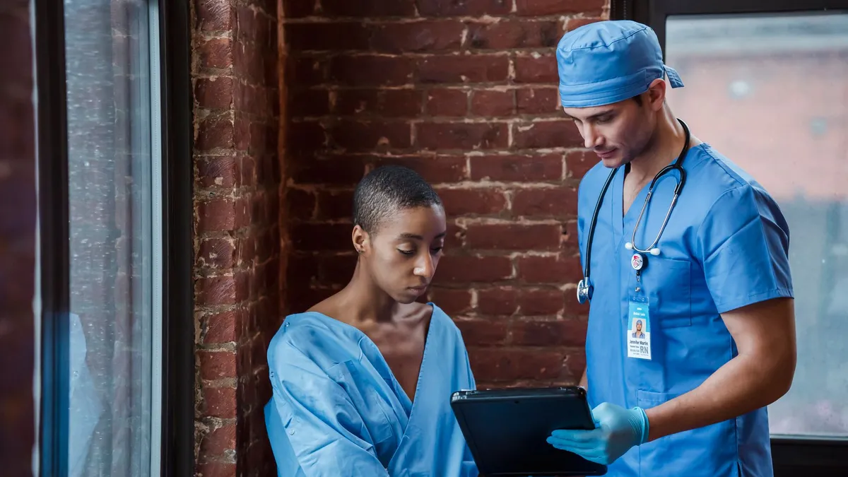 Thoughtful doctor in uniform and gloves showing diagnosis to African American female patient with short hair in blue medical robe in hallway of hospital in daytime