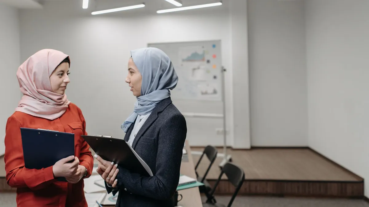 Two professional women in hijabs discussing work while holding clipboards in an office setting.