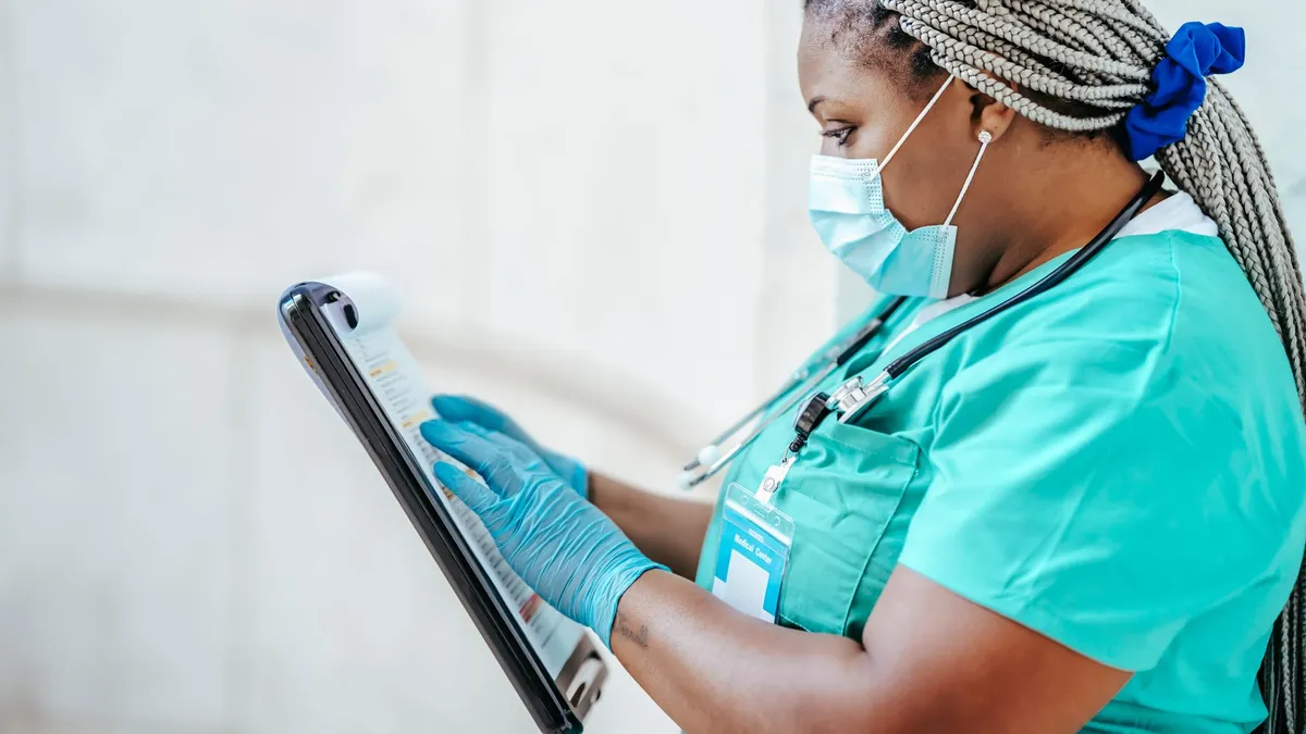 Side view of attentive ethnic female medic with Afro braids in uniform reading paper on clipboard in daytime