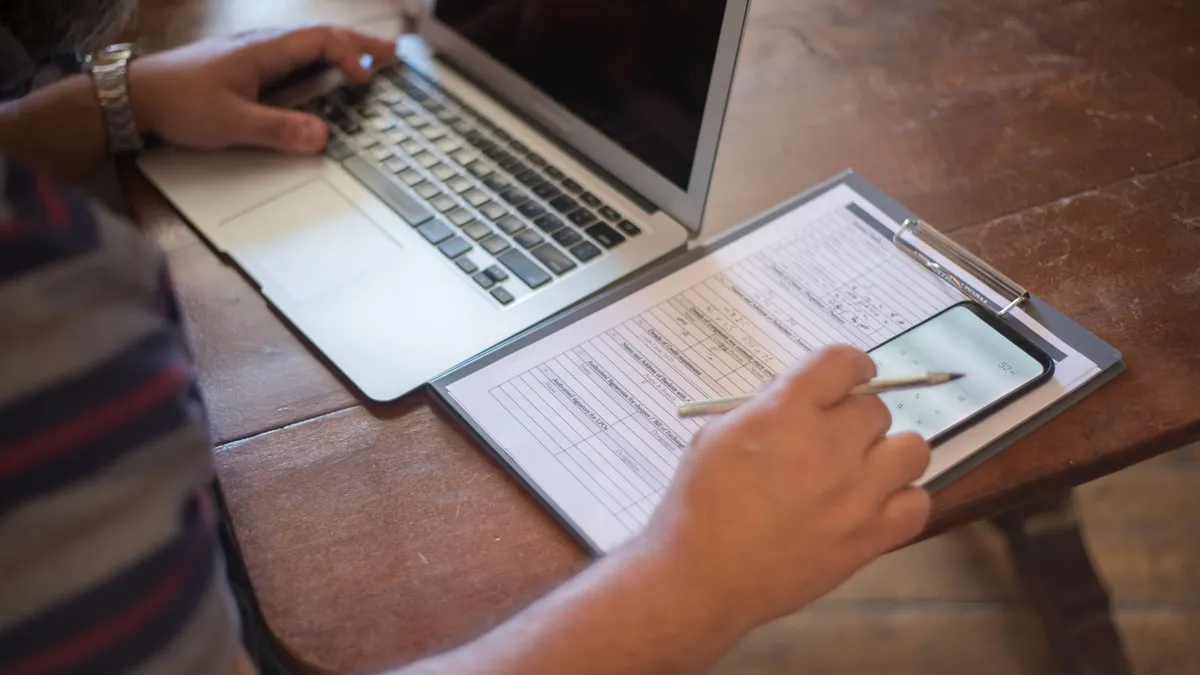Close-up of a person using a laptop and reviewing documents with a smartphone on a wooden table.