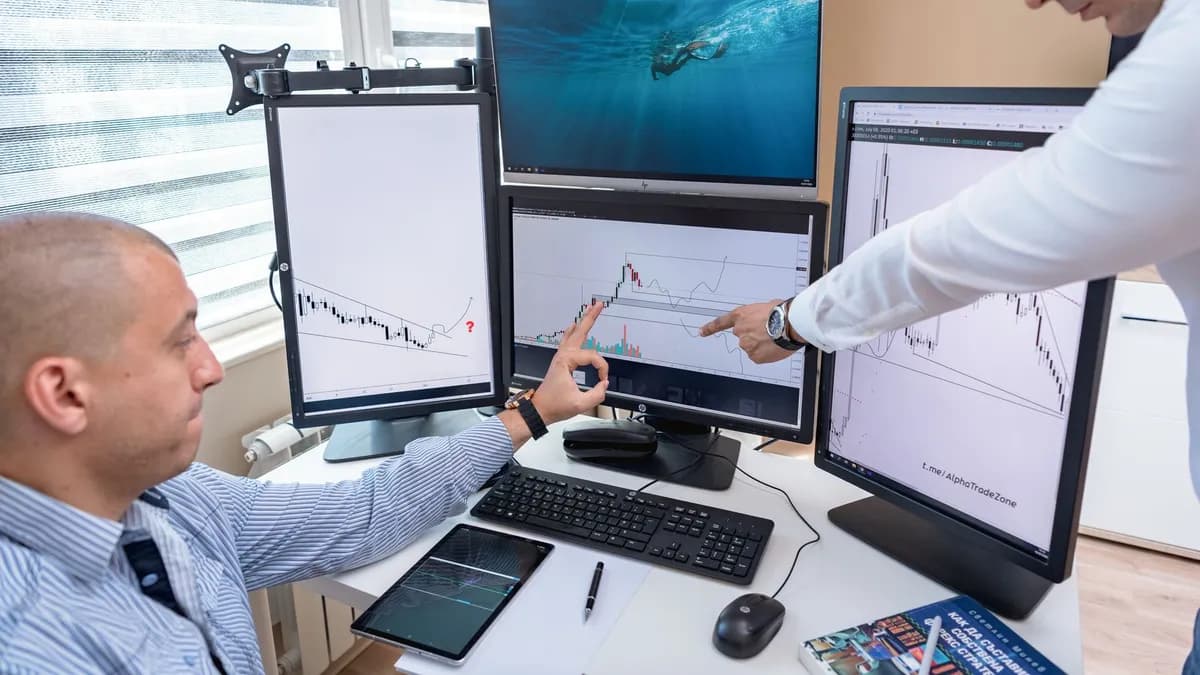 Two business professionals analyzing financial data on multiple computer screens in an office setting.