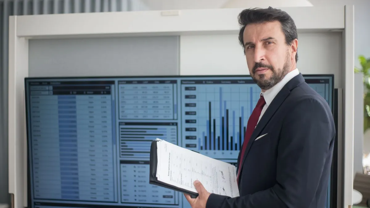 Businessman in a suit holds documents analyzing graphs on a screen, reflecting data analysis.