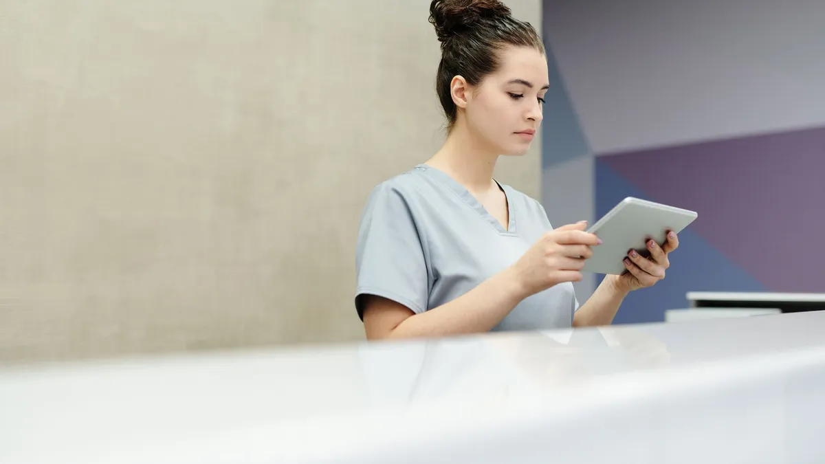 A receptionist in scrubs using a tablet at an office reception desk.
