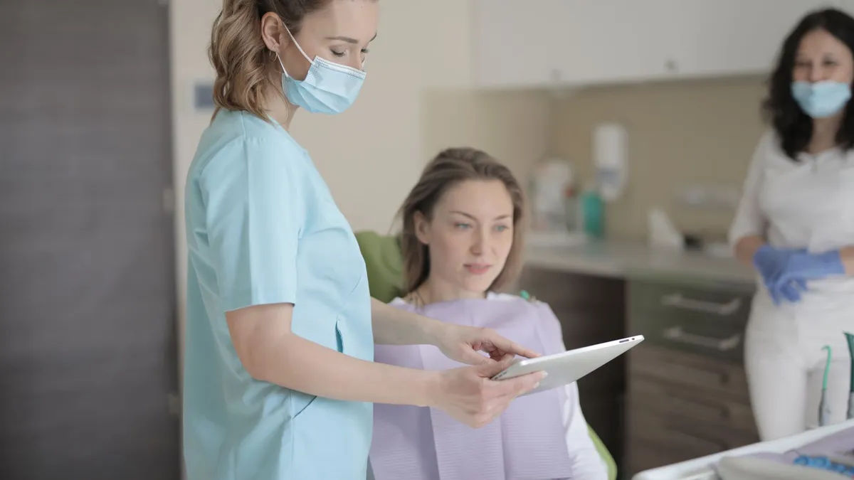 Young female dentist in mask and uniform using tablet while talking with patient before treatment in modern dental clinic in hospital