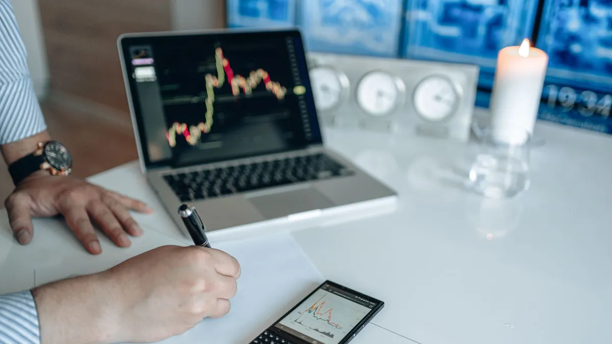 Business analyst writing with laptop and smartphone showing trend graphs on a white desk.