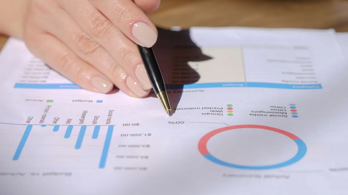 Close-up of a hand with a pen pointing at a financial document with graphs and charts.