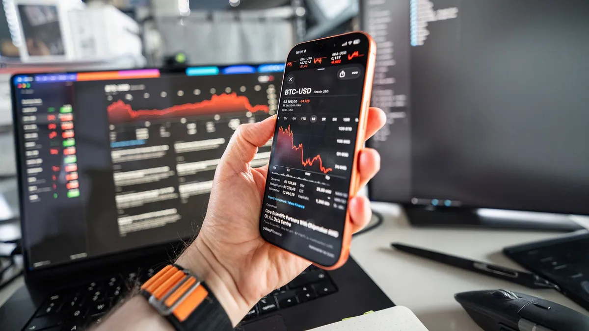 A person analyzing cryptocurrency trends on a smartphone with a declining chart on screen, set against a tech office background.