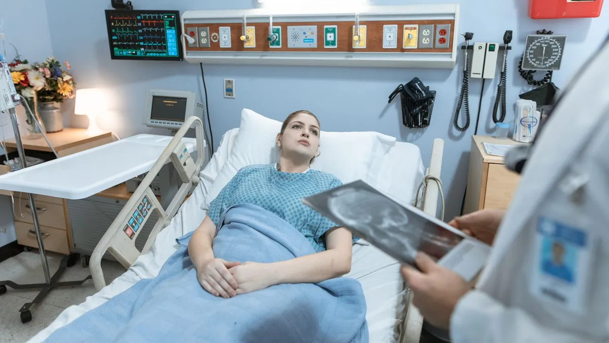 A female patient in a hospital gown receives medical attention from a healthcare professional holding an x-ray in a hospital ward.