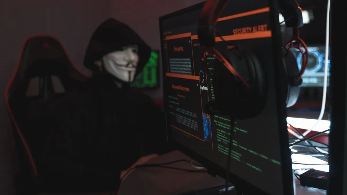 A person wearing a Guy Fawkes mask engaged in hacking activities on a computer in a dimly lit room.