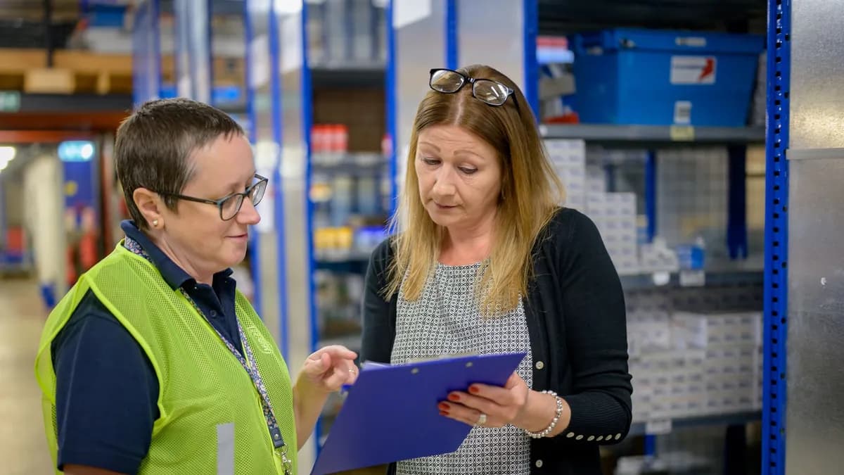 Two women in a warehouse reviewing an inventory checklist on a clipboard.