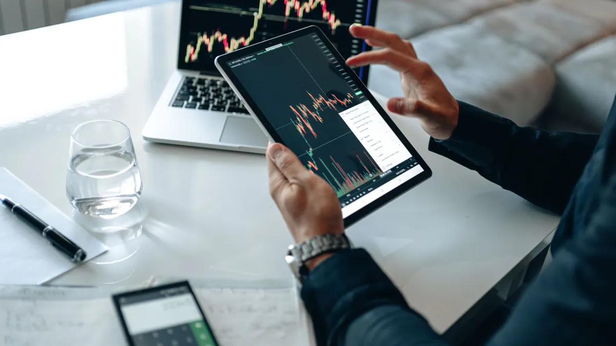 Close-up of a person using a tablet for online trading analysis with a laptop and glass of water nearby.