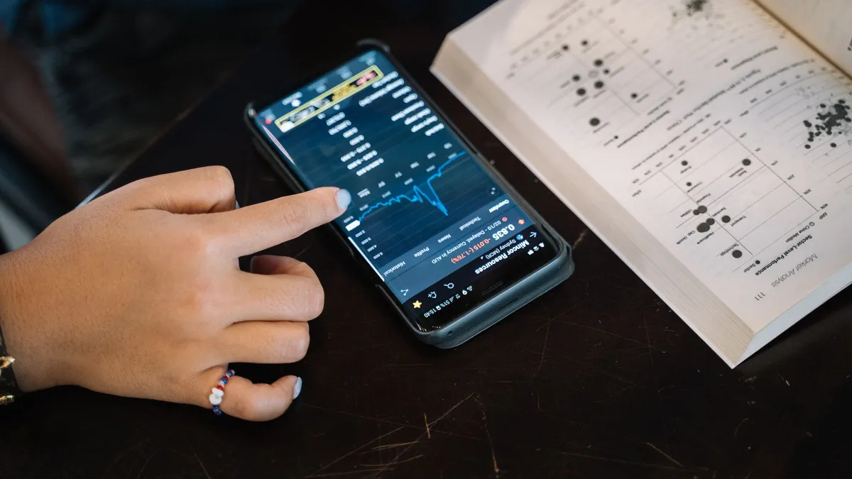 Hand pointing at smartphone displaying financial data, with open book nearby.