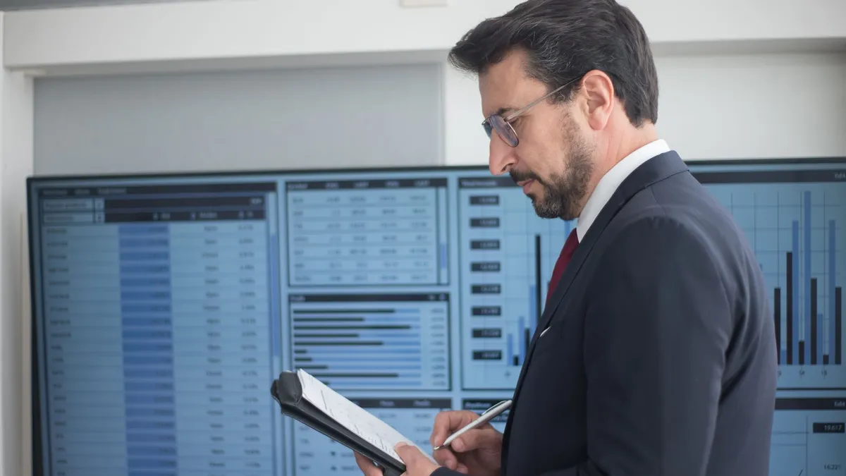 Businessman in a suit analyzing data analytics on large screens, taking notes.