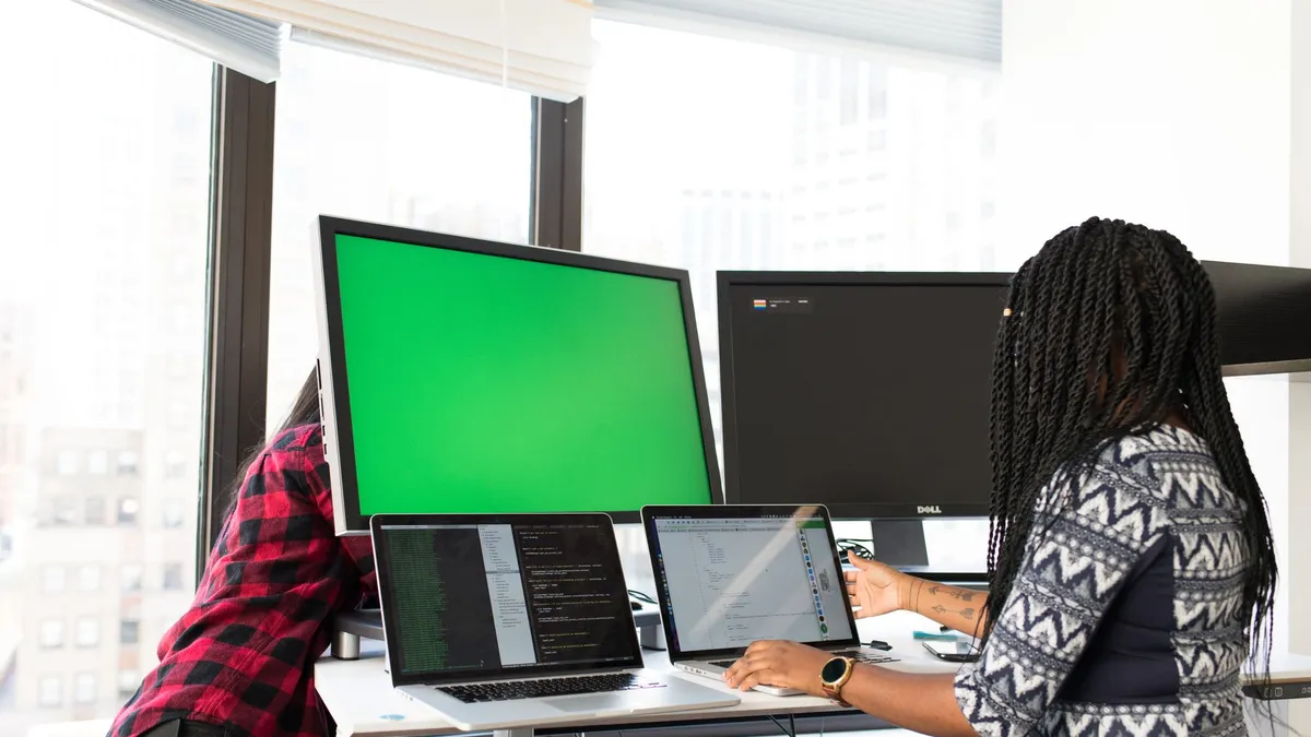 Two women working on laptops and monitors in a bright office setting, focused on technology and teamwork.