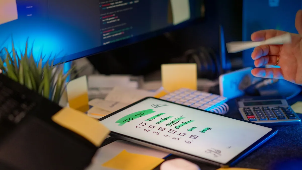 A cluttered desk with a tablet showing a to-do list, monitor showing code, and office supplies.